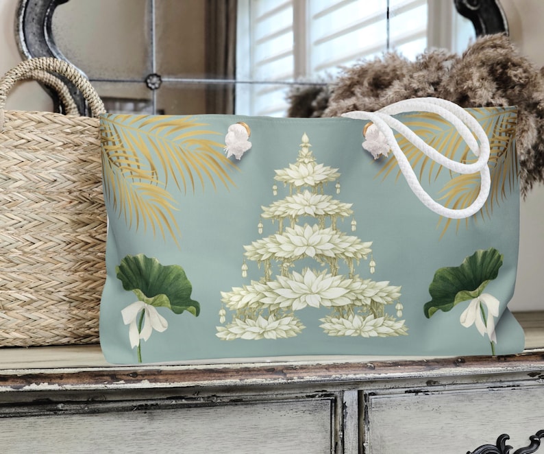 K&ouml;nnte beinhalten: Hellblaue Tragetasche mit wei&szlig;em Seilgriff. Die Tasche hat ein Blumendesign mit wei&szlig;en Lotusbl&uuml;ten und gr&uuml;nen Bl&auml;ttern. Goldene Palmwedel sind ebenfalls auf der Tasche aufgedruckt.