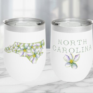 May include: White stainless steel tumbler with a green and white outline of the state of North Carolina with white flowers. The other side of the tumbler has the words "NORTH CAROLINA" in green text with a single white flower.