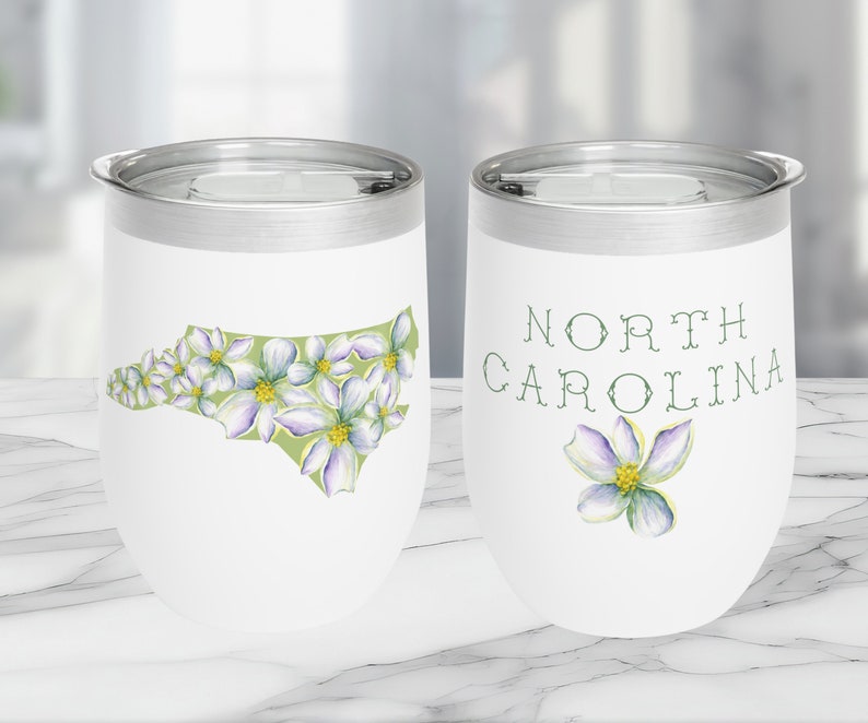 May include: White stainless steel tumbler with a green and white outline of the state of North Carolina with white flowers. The other side of the tumbler has the words "NORTH CAROLINA" in green text with a single white flower.
