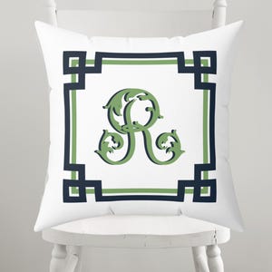May include: White square throw pillow with a green, ornate letter "R" in the center. The letter is framed by a green and navy blue geometric border on a white background. The pillow is on a white chair.