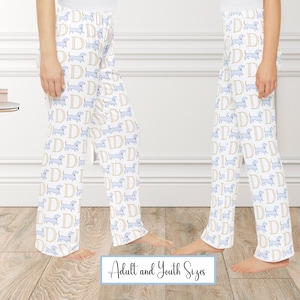 May include: White pajama pants with a repeating pattern of light blue dachshund dogs and the letter "D" in gold. The text "Adult and Youth Sizes" is at the bottom. The pants are shown on two people.