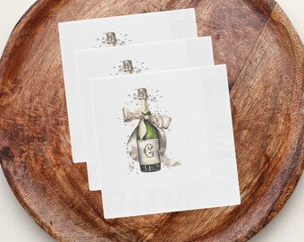 May include: Three white paper napkins with a watercolor illustration of a champagne bottle with a bow and a gold initial. The bottle is surrounded by small silver dots.