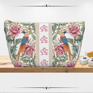 May include: A zippered cosmetic bag with a floral and bird design. The bag features a white background with pink peonies, green foliage, and colorful birds. A central stripe displays the initials "AC".