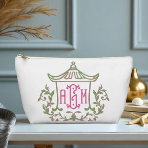 May include: White cosmetic bag with a gold zipper, featuring a green and pink pagoda design with the initials "AGM". The bag is decorated with floral accents and is suitable for storing makeup and toiletries.