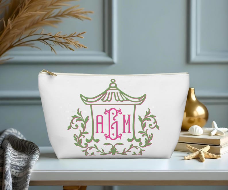 May include: White cosmetic bag with a gold zipper, featuring a green and pink pagoda design with the initials "AGM". The bag is decorated with floral accents and is suitable for storing makeup and toiletries.