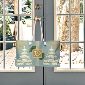 May include: A light blue tote bag with a floral design and a gold monogram. The bag features white rope handles and is hanging on a door. The design includes gold accents and a repeating pattern.