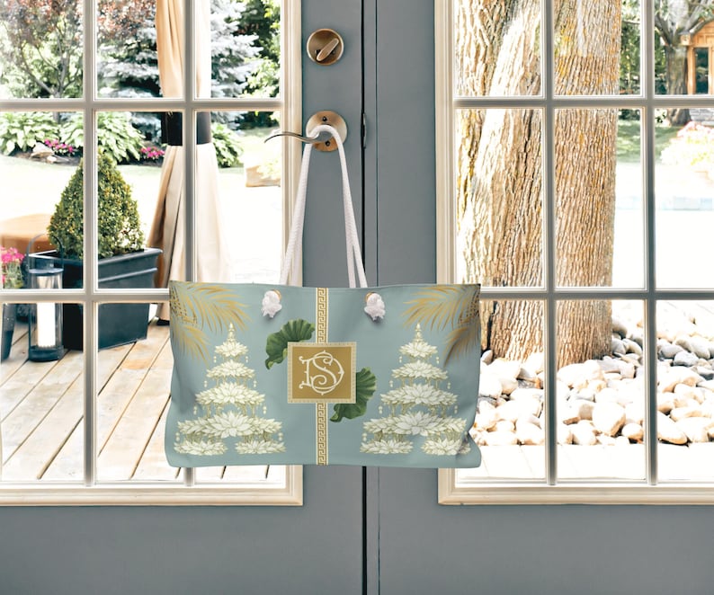 May include: A light blue tote bag with a floral design and a gold monogram. The bag features white rope handles and is hanging on a door. The design includes gold accents and a repeating pattern.
