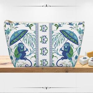 May include: A white cosmetic bag with a blue and green monkey and umbrella pattern. The bag has a gold zipper and a central panel with the letter "E" repeated. The design includes floral and geometric elements.