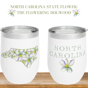 May include: Two white stainless steel tumblers with lids. One tumbler features a green outline of the state of North Carolina filled with white dogwood flowers. The other tumbler has the words "NORTH CAROLINA" in green script with a single white dogwood flower.