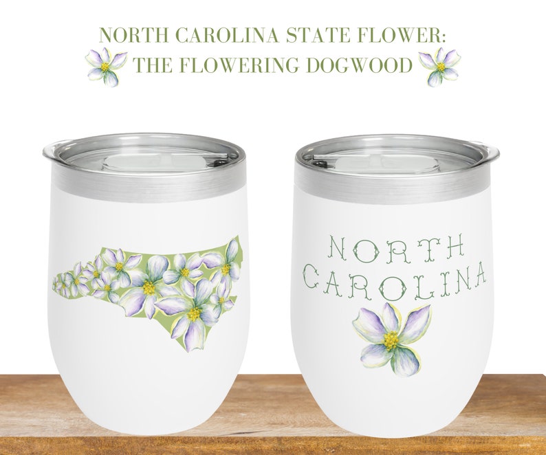 May include: Two white stainless steel tumblers with lids. One tumbler features a green outline of the state of North Carolina filled with white dogwood flowers. The other tumbler has the words "NORTH CAROLINA" in green script with a single white dogwood flower.