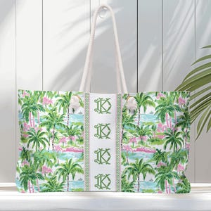 May include: A large tote bag with a tropical print featuring palm trees, pink flamingos, and a blue ocean scene. The bag has white rope handles and a white center stripe with green monogrammed initials. The bag is made of a durable fabric.