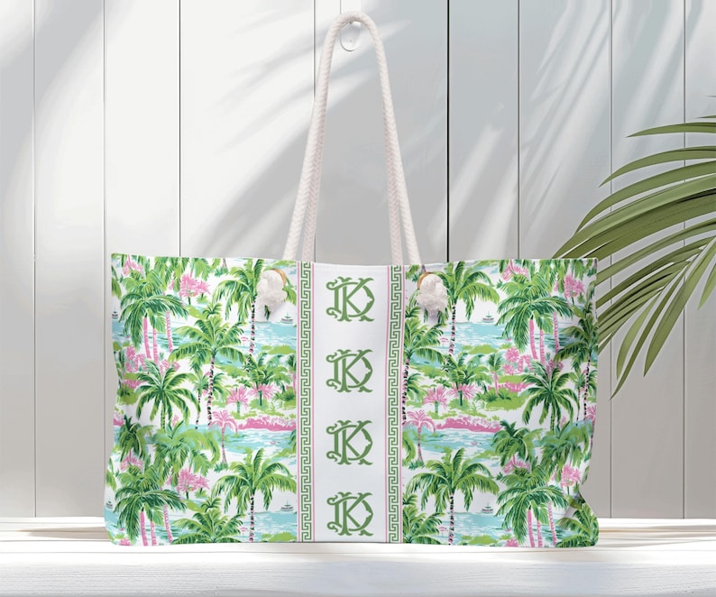 May include: A large tote bag with a tropical print featuring palm trees, pink flamingos, and a blue ocean scene. The bag has white rope handles and a white center stripe with green monogrammed initials. The bag is made of a durable fabric.