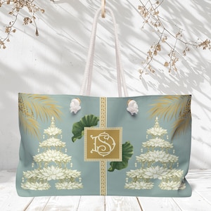May include: Large tote bag with a light blue background featuring a floral design with white and gold accents. The bag has white rope handles and a gold monogram. The design includes lotus flowers and palm fronds.