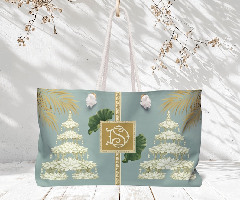 May include: Large tote bag with a light blue background featuring a floral design with white and gold accents. The bag has white rope handles and a gold monogram. The design includes lotus flowers and palm fronds.
