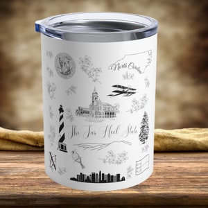 May include: White stainless steel tumbler with a black and white design featuring various illustrations of North Carolina landmarks, including a lighthouse, a building, a plane, and a pine tree. The text "The Tar Heel State" is printed on the tumbler.