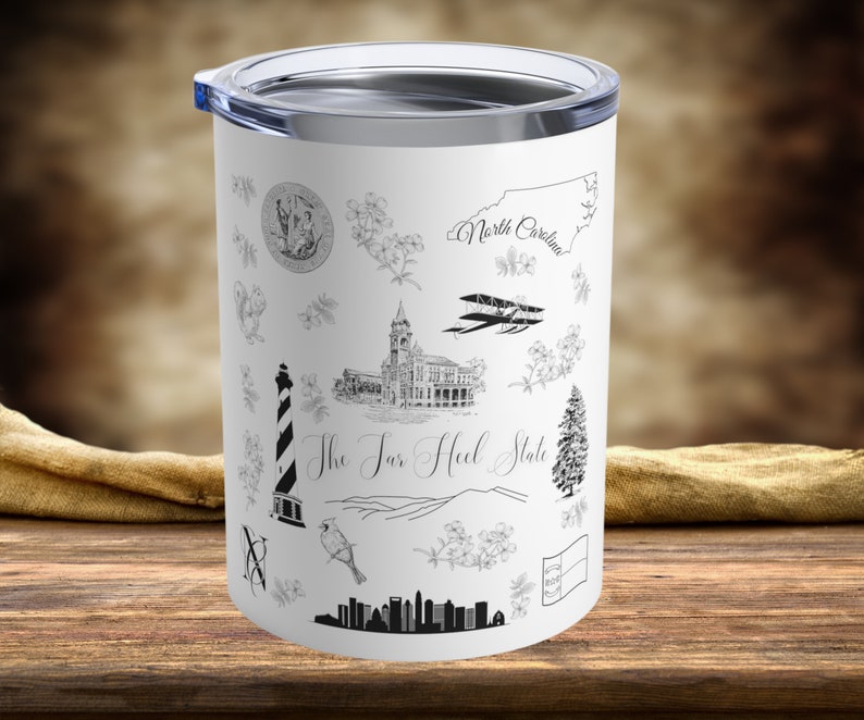 May include: White stainless steel tumbler with a black and white design featuring various illustrations of North Carolina landmarks, including a lighthouse, a building, a plane, and a pine tree. The text "The Tar Heel State" is printed on the tumbler.