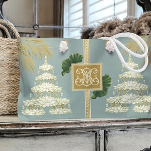 May include: A large tote bag with a light blue background featuring a pagoda design with white lotus flowers and gold accents. The bag has white rope handles and a gold monogram. A woven basket is visible on the left.