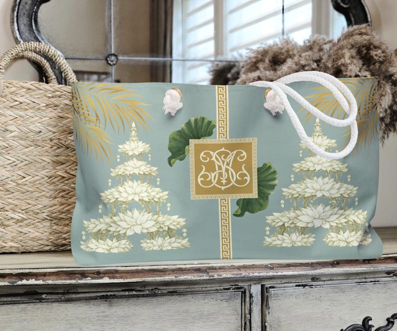 May include: A large tote bag with a light blue background featuring a pagoda design with white lotus flowers and gold accents. The bag has white rope handles and a gold monogram. A woven basket is visible on the left.