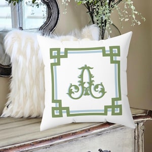 Peut inclure: Coussin carr&eacute; blanc avec une bordure g&eacute;om&eacute;trique verte et bleu clair et un "A" vert orn&eacute; au centre. Le coussin est pos&eacute; sur une surface en bois blanc vieilli.