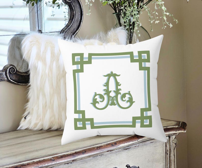 Peut inclure: Coussin carr&eacute; blanc avec une bordure g&eacute;om&eacute;trique verte et bleu clair et un "A" vert orn&eacute; au centre. Le coussin est pos&eacute; sur une surface en bois blanc vieilli.