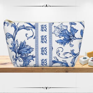 May include: A white cosmetic bag with a blue floral design and gold zipper. The bag features a repeating pattern of stylized flowers and leaves. The bag has a rectangular shape and is sitting on a wooden shelf.