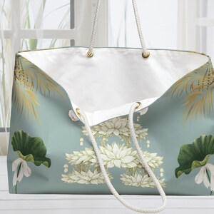K&ouml;nnte beinhalten: Gro&szlig;e Tragetasche mit hellt&uuml;rkisfarbenem Hintergrund und floralem Design. Die Tasche hat wei&szlig;e Lotusblumen, gr&uuml;ne Seerosenbl&auml;tter und goldene Palmfarnwedel. Sie hat wei&szlig;e Seilgriffe und ein wei&szlig;es Innenfutter.