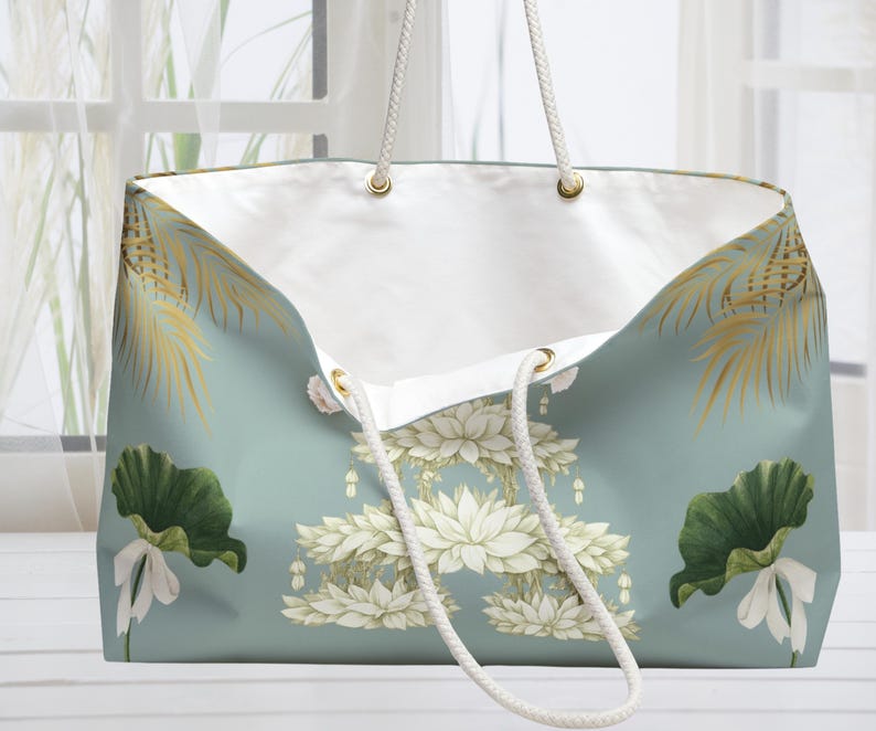 K&ouml;nnte beinhalten: Gro&szlig;e Tragetasche mit hellt&uuml;rkisfarbenem Hintergrund und floralem Design. Die Tasche hat wei&szlig;e Lotusblumen, gr&uuml;ne Seerosenbl&auml;tter und goldene Palmfarnwedel. Sie hat wei&szlig;e Seilgriffe und ein wei&szlig;es Innenfutter.
