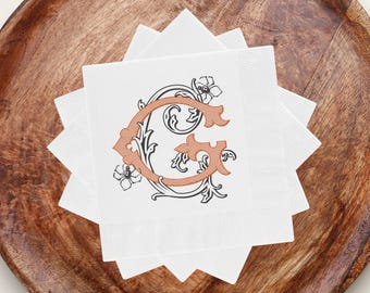 May include: White square napkins with a decorative letter "G" in orange and black, featuring floral accents. The napkins are stacked on a wooden tray, suitable for a party or event.