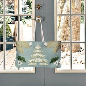 K&ouml;nnte beinhalten: Eine hellblaue Tragetasche mit floralem Design, die an einem T&uuml;rgriff h&auml;ngt. Die Tasche zeigt eine gestufte Blumenanordnung mit wei&szlig;en und goldenen Akzenten sowie gr&uuml;ne Bl&auml;tter und Palmwedel. Die wei&szlig;en Tr&auml;ger der Tasche sind sichtbar.
