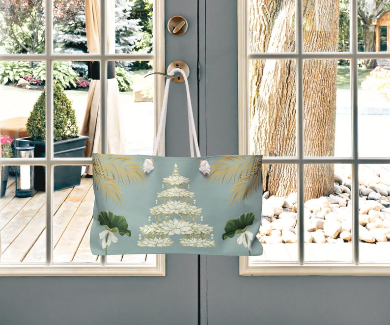 K&ouml;nnte beinhalten: Eine hellblaue Tragetasche mit floralem Design, die an einem T&uuml;rgriff h&auml;ngt. Die Tasche zeigt eine gestufte Blumenanordnung mit wei&szlig;en und goldenen Akzenten sowie gr&uuml;ne Bl&auml;tter und Palmwedel. Die wei&szlig;en Tr&auml;ger der Tasche sind sichtbar.