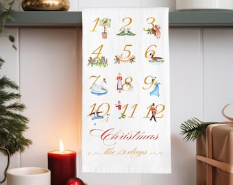 May include: A white kitchen towel with a gold design of the 12 days of Christmas. The numbers 1 through 12 are displayed with illustrations of each day's gift. The text "Christmas the 12 days" is written in cursive.