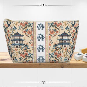 May include: A beige cosmetic bag with a gold zipper, featuring a floral and bird pattern with pagodas. A central white stripe displays the letter "A" repeated. Mahjong tiles are visible on the wooden surface.