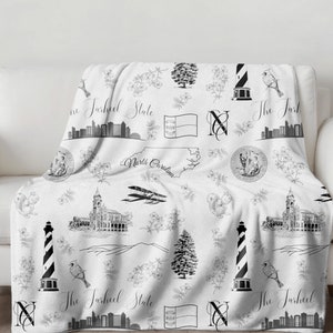 Peut inclure: Jet&eacute; de canap&eacute; noir et blanc avec un motif r&eacute;p&eacute;t&eacute; de monuments de Caroline du Nord, notamment le b&acirc;timent du Capitole de l'&Eacute;tat, un phare et le drapeau de l'&Eacute;tat. Le texte "The Tarheel State" est r&eacute;p&eacute;t&eacute; dans tout le motif.