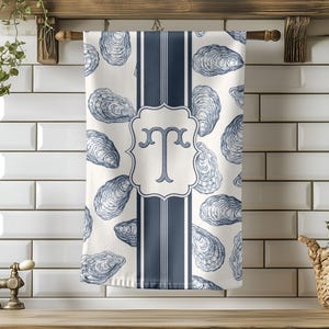 May include: A white kitchen towel with a blue oyster shell pattern and a navy blue stripe. The towel features a decorative monogram letter "T" in a framed design. The towel is hanging on a wooden rod.