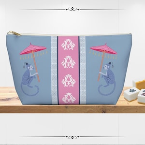 May include: A light blue cosmetic bag with a gold zipper. The bag features a pink and white stripe down the center with the letter "A" repeated. Two blue monkeys holding pink umbrellas are on the front.