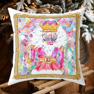 May include: A white throw pillow with a gold frame featuring a colorful painting of a Nutcracker. The Nutcracker is wearing a red and gold costume with a crown and holding a staff. The background is a colorful abstract design.
