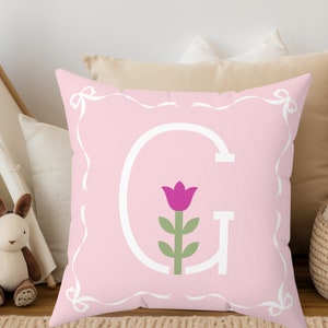 May include: Pink pillow with a white border and a white letter G with a pink flower inside. The pillow has a white ribbon design.