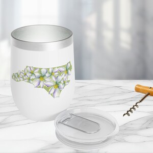 May include: White stainless steel tumbler with a green outline of the state of North Carolina, decorated with white flowers. The tumbler has a silver lid with a built-in bottle opener.