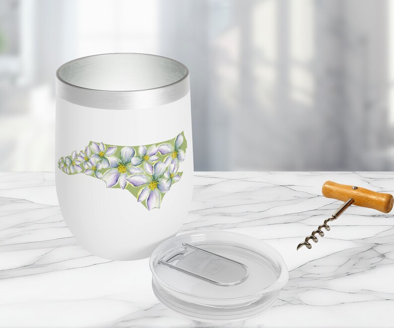May include: White stainless steel tumbler with a green outline of the state of North Carolina, decorated with white flowers. The tumbler has a silver lid with a built-in bottle opener.