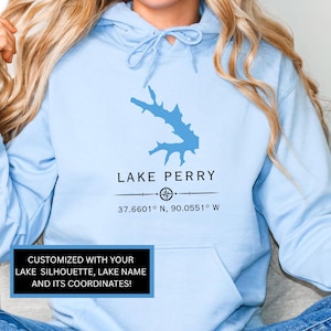 May include: Light blue hoodie featuring a blue lake silhouette, the words "LAKE PERRY", and coordinates. The text "CUSTOMIZED WITH YOUR LAKE SILHOUETTE, LAKE NAME AND ITS COORDINATES!" is displayed in a black box.