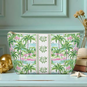 May include: A white cosmetic bag with a tropical palm tree and ocean scene print. The bag features a gold zipper and a green and white Greek key design. The bag is approximately 10 inches wide and 6 inches tall.