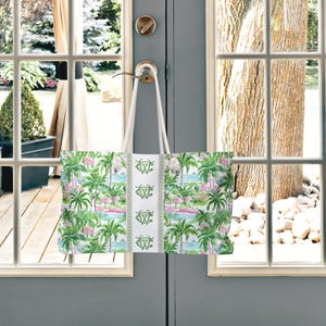 May include: A large tote bag with a tropical print of palm trees, flamingos, and water scenes in green, pink, and blue. The bag has white handles and a white center panel with a green design. The bag is hanging on a door handle.