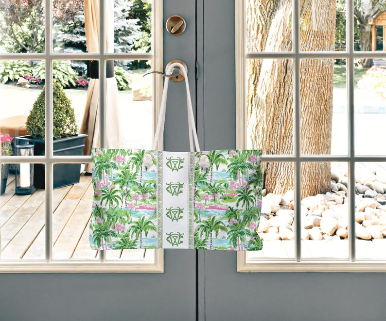 May include: A large tote bag with a tropical print of palm trees, flamingos, and water scenes in green, pink, and blue. The bag has white handles and a white center panel with a green design. The bag is hanging on a door handle.