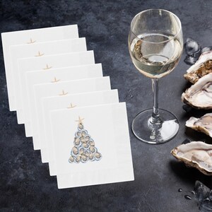 May include: A stack of eight white napkins with a blue and white oyster shell Christmas tree design. The napkins are on a dark gray surface with a glass of white wine and oyster shells.