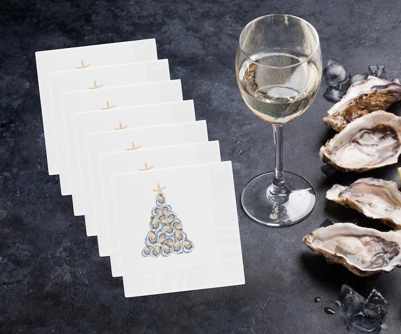 May include: A stack of eight white napkins with a blue and white oyster shell Christmas tree design. The napkins are on a dark gray surface with a glass of white wine and oyster shells.