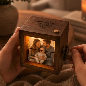 May include: A square wooden music box with a dark brown finish featuring an internal illuminated photo of a family. The top is engraved with the text I love you, baby. 24.07.2026 and two small hearts. A small brass button is on the top right corner.