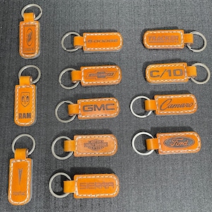 Op de afbeelding: Een verzameling van 11 leren sleutelhangers met verschillende logo's en merknamen, waaronder Buick, Dodge, Tracker, C/10, Ram, GMC, Harley Davidson, Sierra, Camaro, Ford en Pontiac. De sleutelhangers zijn bruin en hebben een gestikte rand.