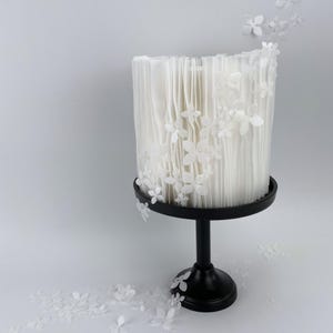 Op de afbeelding: Een witte taart met een textuur, verticaal ontwerp en witte bloemaccenten. De taart staat op een zwarte, ronde taartstandaard met een enkele steel. Witte bloemdecoraties zijn verspreid.