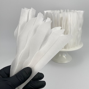 Könnte beinhalten: Eine Hand mit schwarzem Handschuh hält ein Bündel weißer, wellenförmiger Papierstreifen. Im Hintergrund zeigt ein weißer Kuchenständer eine zylindrische Anordnung ähnlicher Papierstreifen, die einen geschichteten Effekt erzeugt. Die Gesamtästhetik ist sauber und minimalistisch.