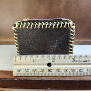 K&ouml;nnte beinhalten: Eine dunkelbraune Lederbox mit sichtbaren cremefarbenen N&auml;hten im Baseball-Stil entlang der Kanten. Ein Fiskars-Lineal liegt davor und zeigt, dass die Box eine Breite von etwa 8,25 cm hat. Die Oberseite ist mit einem dunkelvioletten Lederpaneel versehen.